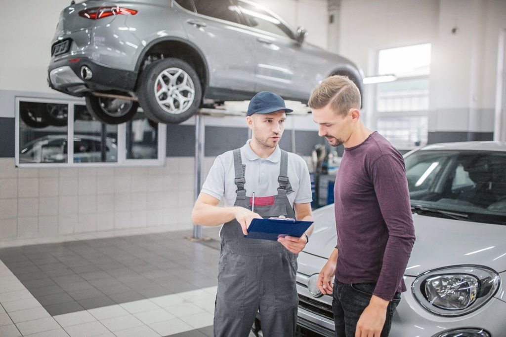A mechanic reviews a report with a client at the best car inspection service Houston, with a lifted car in the background.
