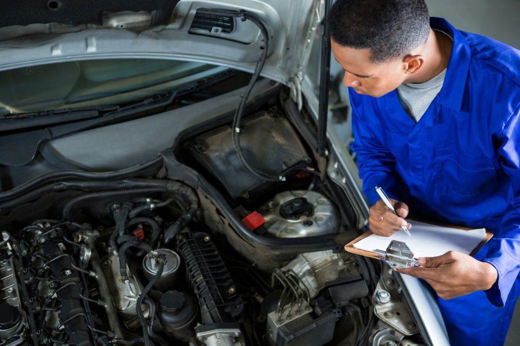 A mechanic in blue uniform conducting a third party inspection Houston on a car engine with a clipboard.