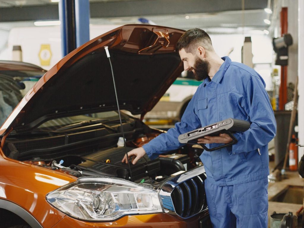 Mechanic using diagnostic tool for used auto inspection Houston on an orange car's open hood.