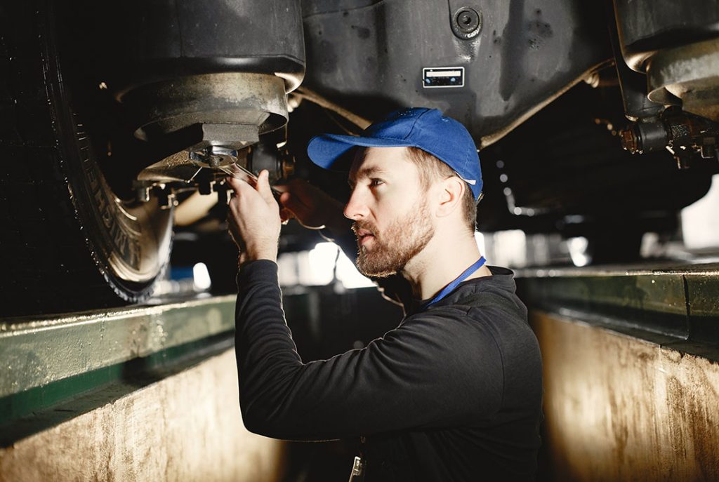 Certified third party car inspector performing a thorough undercarriage inspection and adjustment in a service bay.