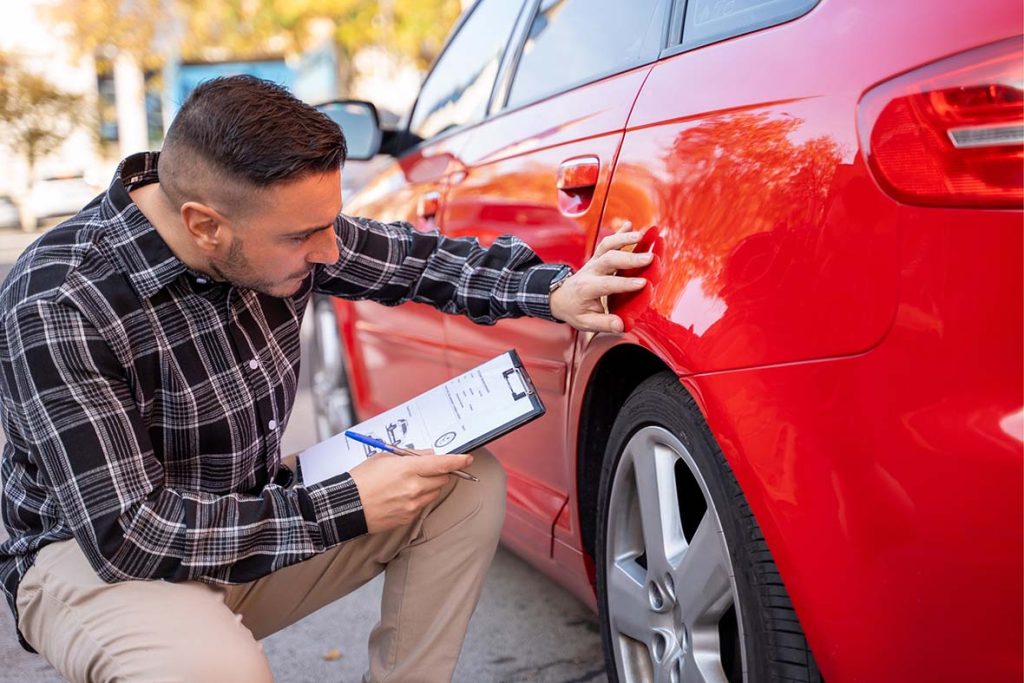 Expert performing third party car inspection with report; checking the red car's body panel for exterior damage.